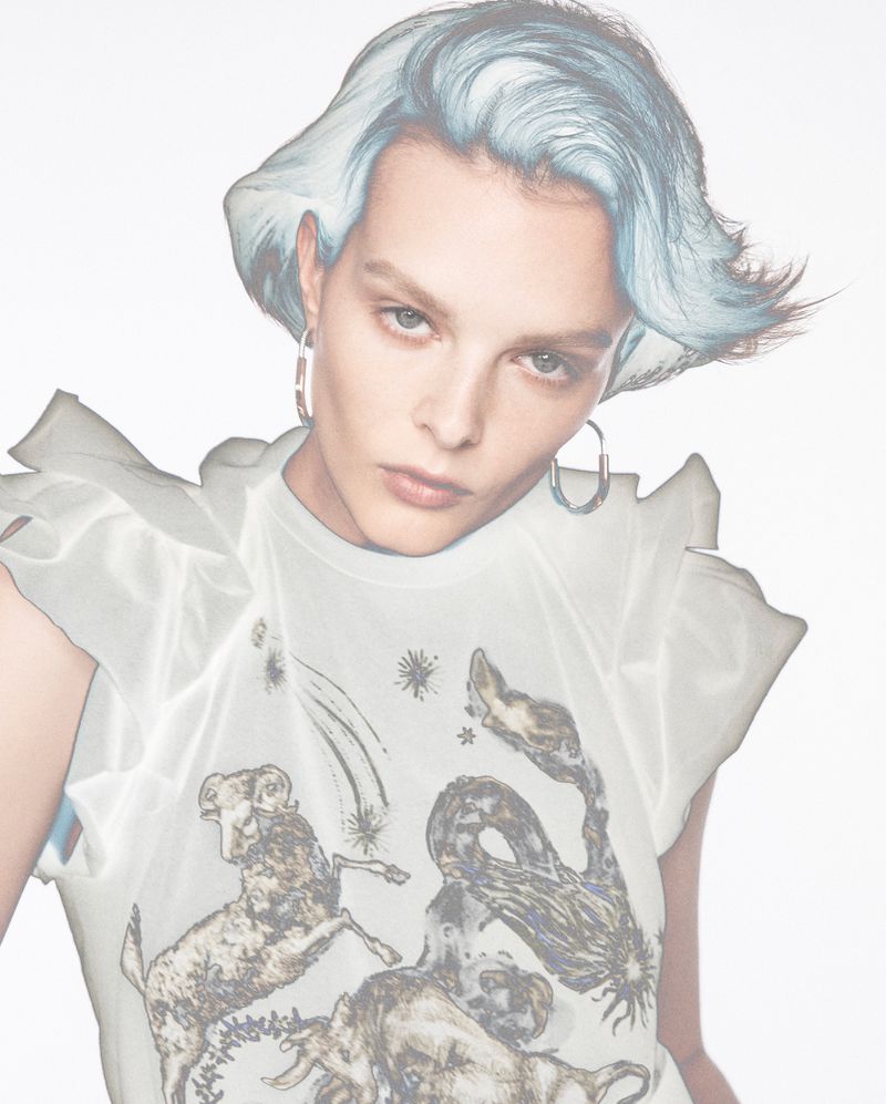 Close-up studio portrait of an adult female model with light blue hair, hoop earrings, and white t-shirt with animal graphic print.