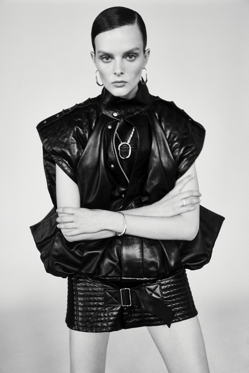 Monochrome portrait of a woman with slicked-back hair in black leather jacket and skirt against a pale background in photo studio lighting.
