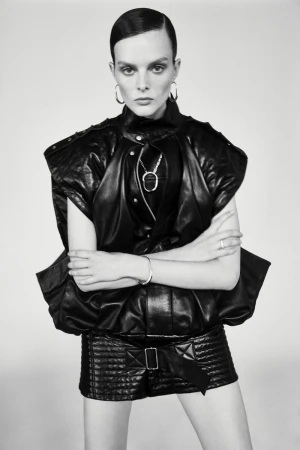 Monochrome portrait of a woman with slicked-back hair in black leather jacket and skirt against a pale background in photo studio lighting.