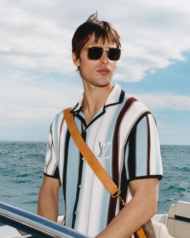 Stylish adult male in sunglasses on a boat, wearing a striped shirt with a shoulder bag, against a sky and ocean backdrop.