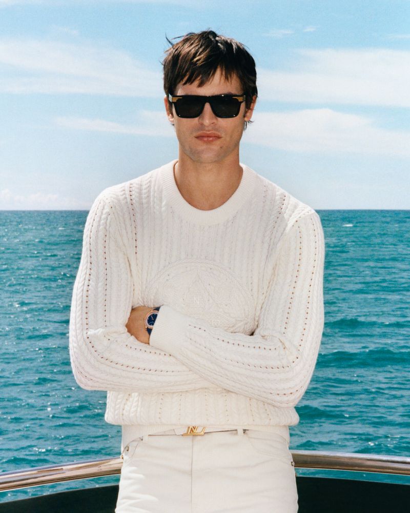 Adult male stands confidently on a boat, wearing a white sweater, white pants, and sunglasses, with a blue sea and sky background.