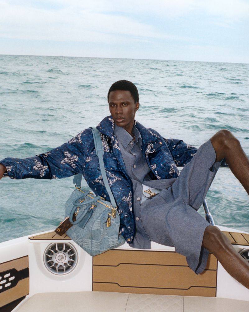 A stylish man in a blue patterned jacket and shorts poses on a white boat, accessorized with a blue satchel, against a backdrop of the ocean.