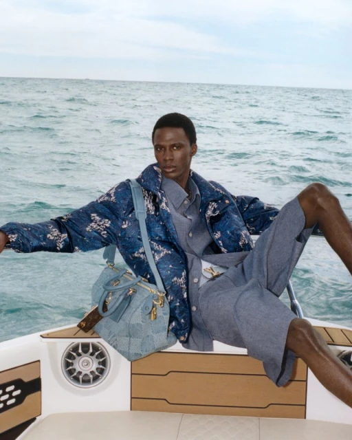A stylish man in a blue patterned jacket and shorts poses on a white boat, accessorized with a blue satchel, against a backdrop of the ocean.