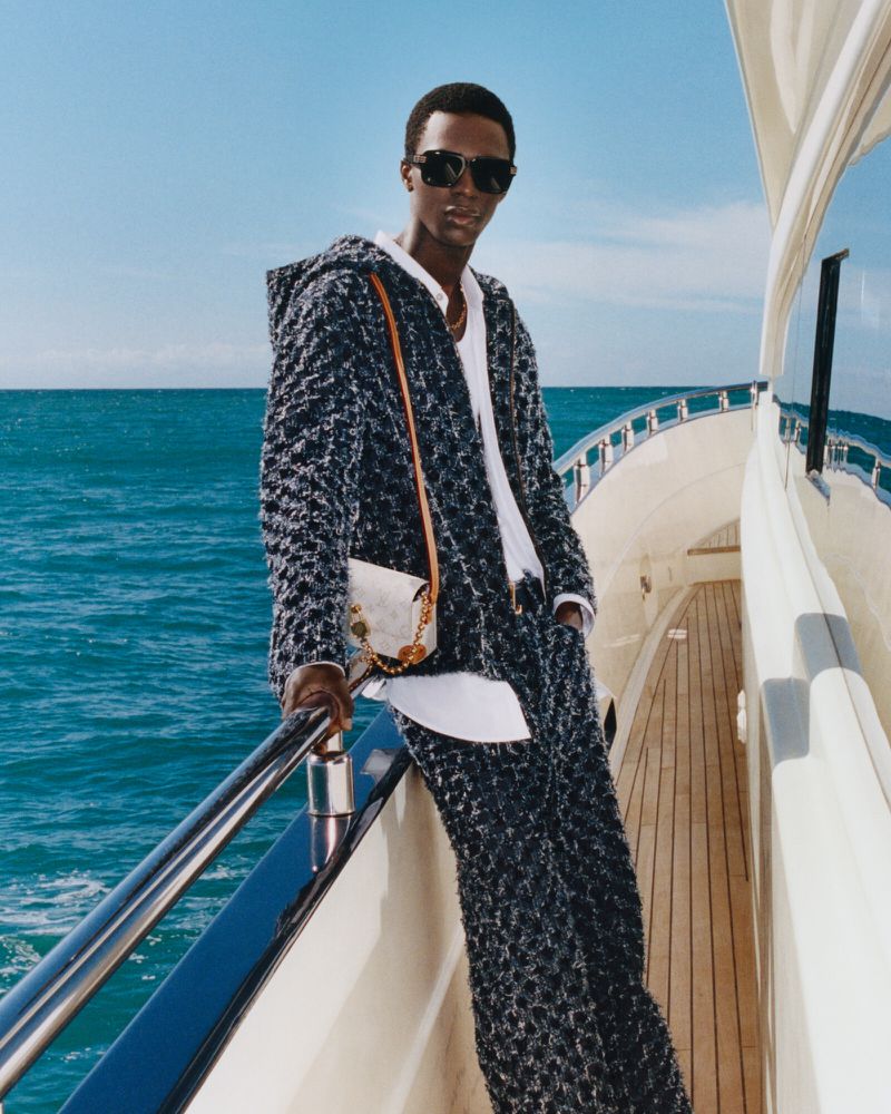 An African-American man in stylish hooded jacket, matching pants, sunglasses, and white shirt with a purse, poses on a yacht.