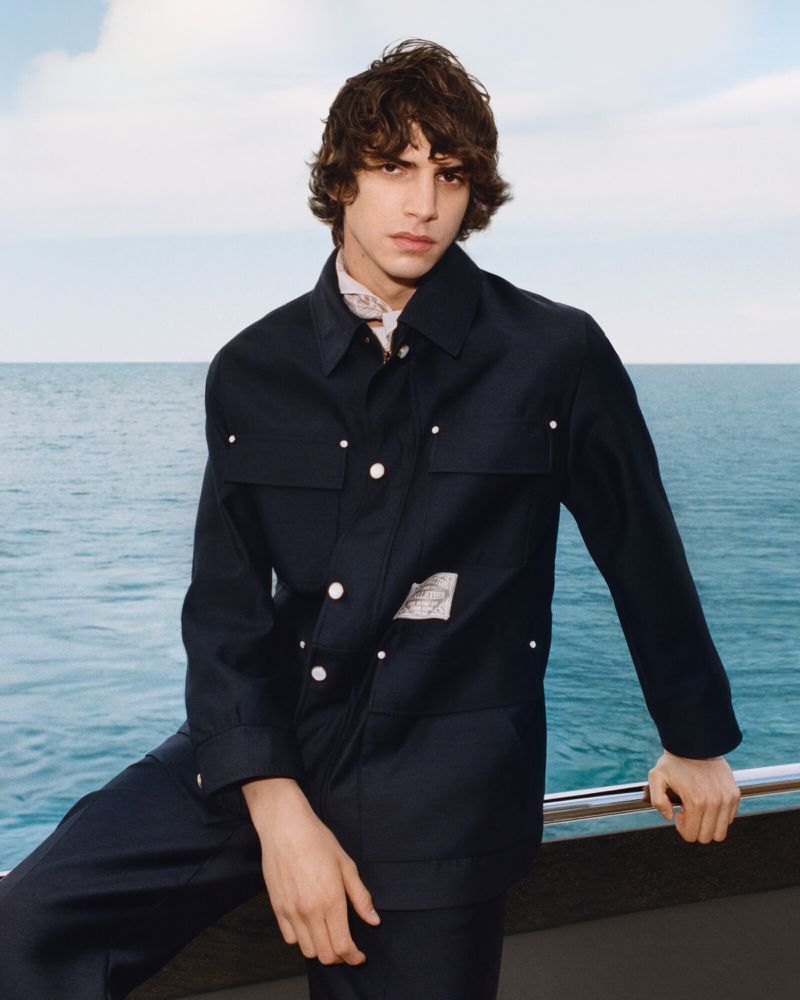 Model wearing a dark jacket standing outdoors against a backdrop of open ocean and a light sky