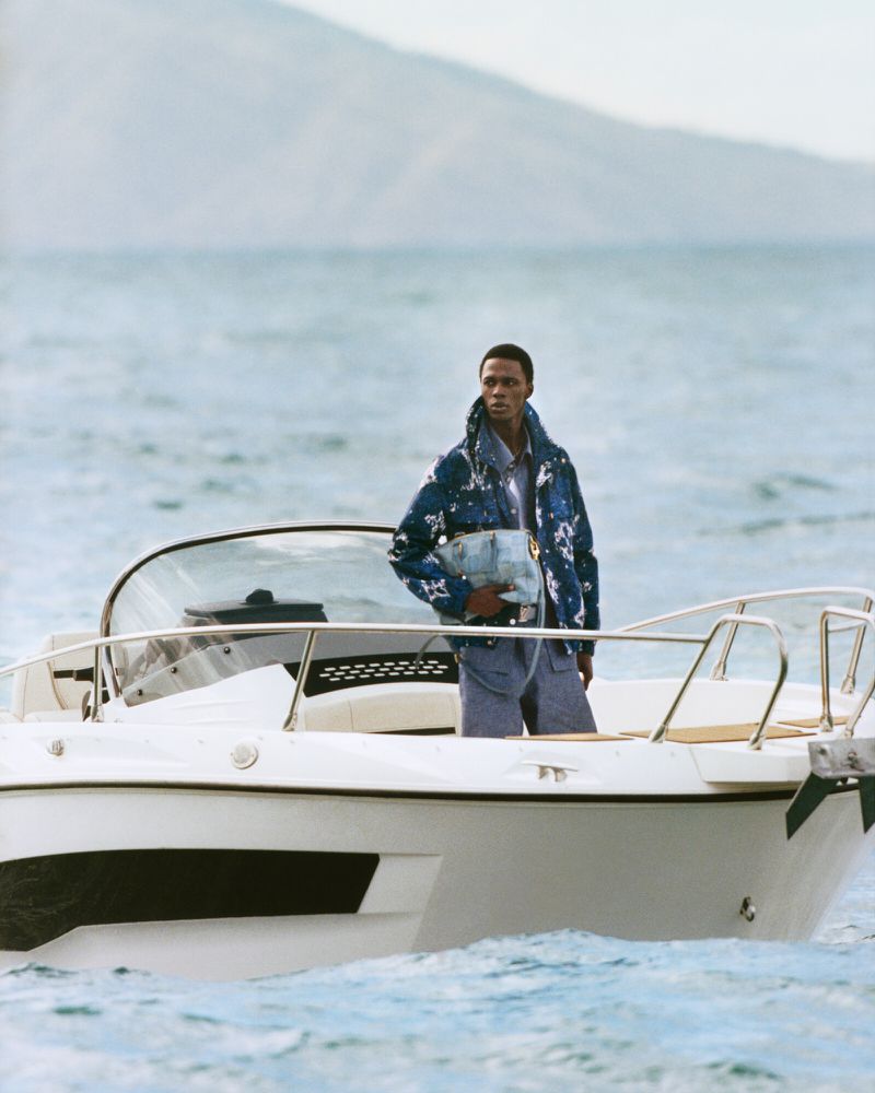 An African American man poses on a white motorboat wearing a patterned jacket and blue shorts on blue wavy sea with a mountain in the distance.