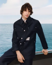 Model wearing a dark jacket standing outdoors against a backdrop of open ocean and a light sky