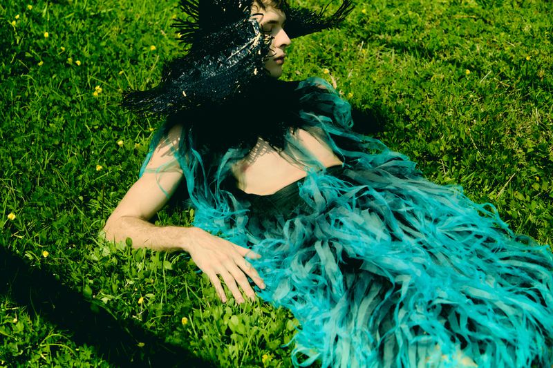 Person in elaborate feathered headpiece and turquoise tattered dress lying on grass in natural daylight.