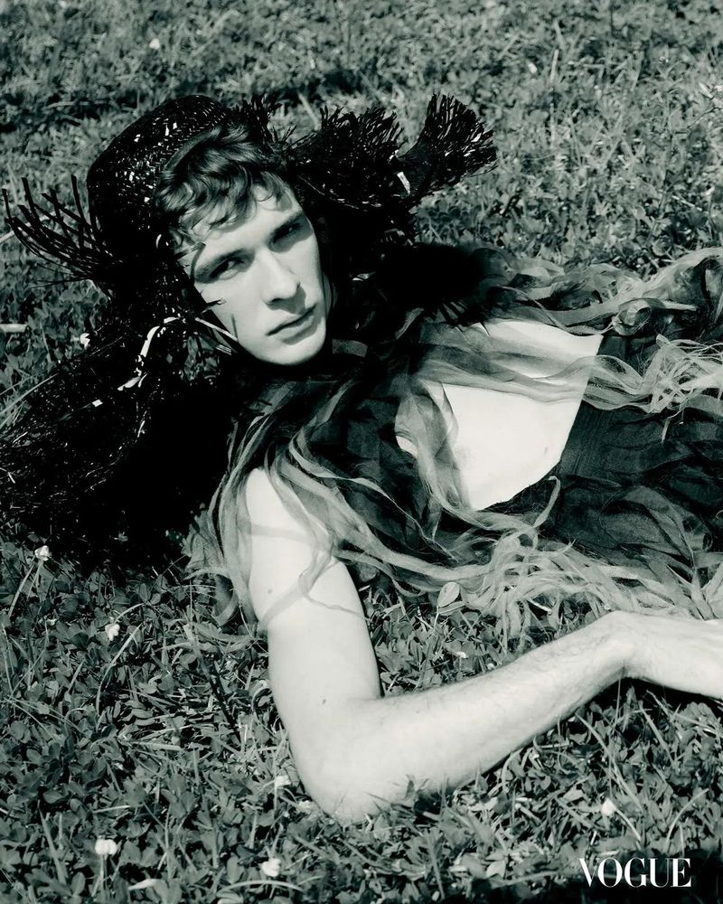 A male model with curly hair lies on a grassy field wearing a black hat and a sheer, flowing top, set against a soft, natural backdrop.