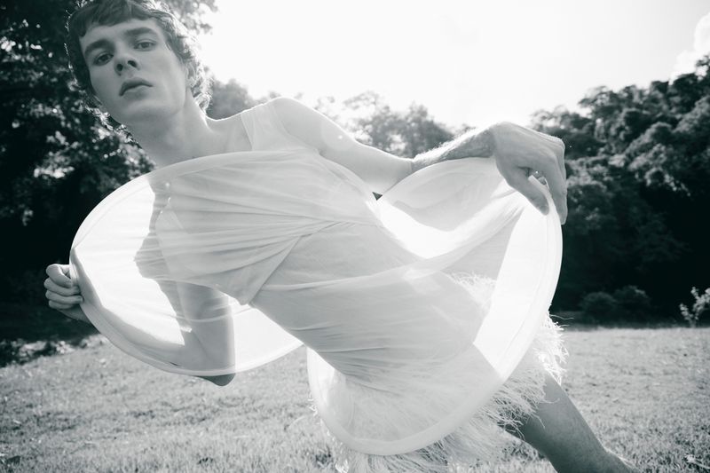 Monochromatic portrait of a slender adult male in sheer fabric and feather details, posing in a sunlit grassy field with tree background.