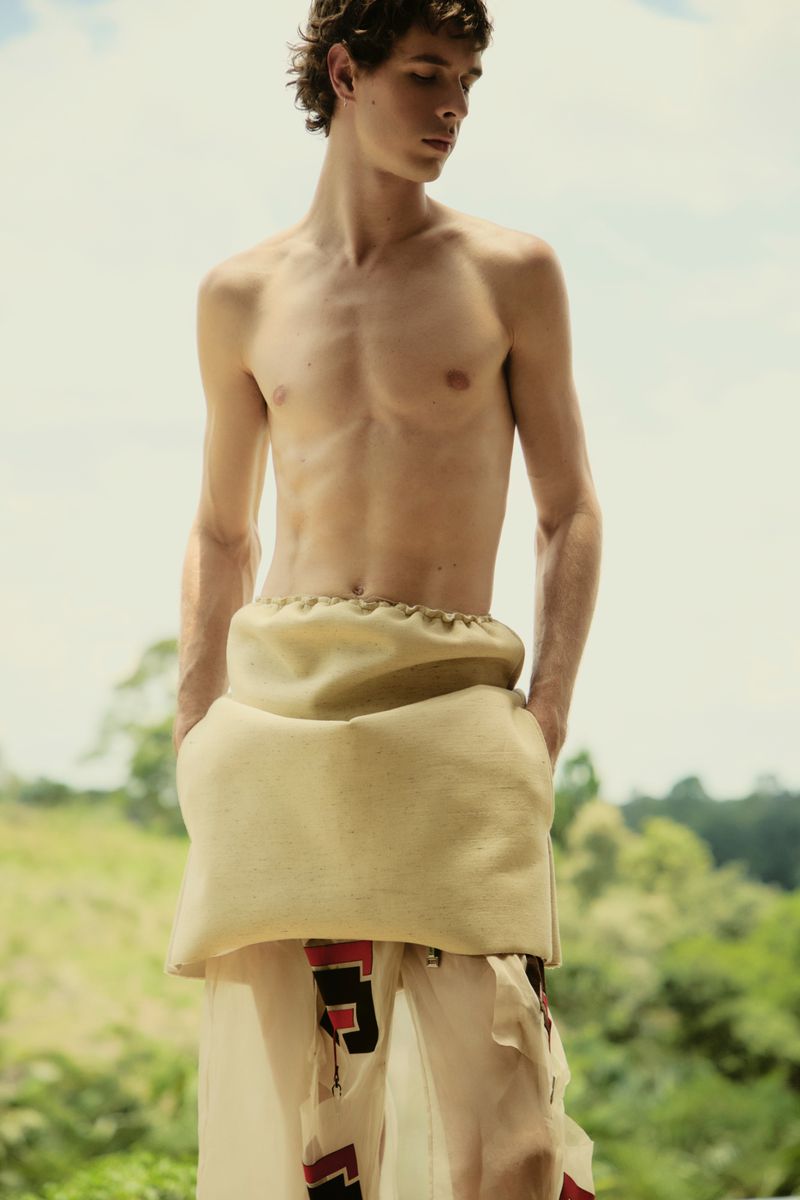 Young male fashion model poses outdoors wearing unique beige fabric wrap and patterned pants against a blurred natural background.