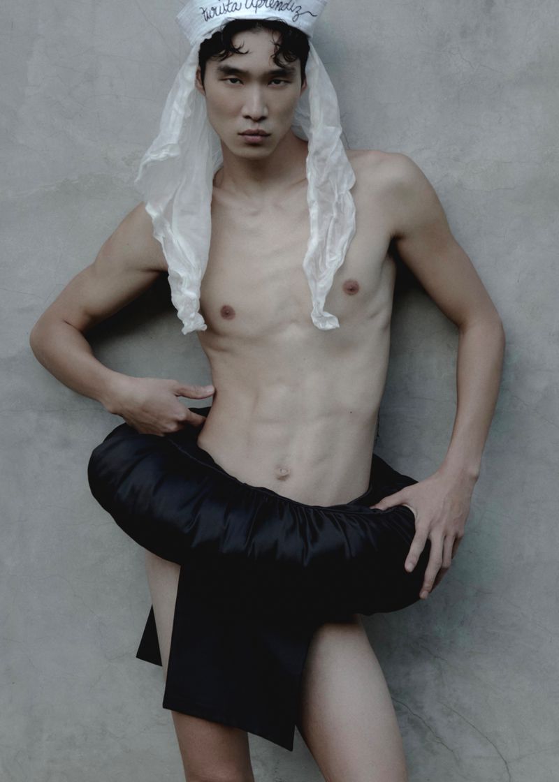 Shirtless Asian man with toned physique poses in front of grey wall wearing a white hat and black skirt-like garment.