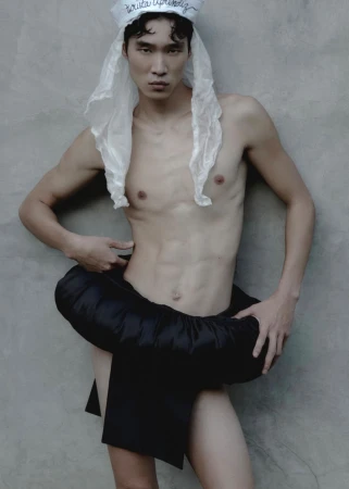 Shirtless Asian man with toned physique poses in front of grey wall wearing a white hat and black skirt-like garment.