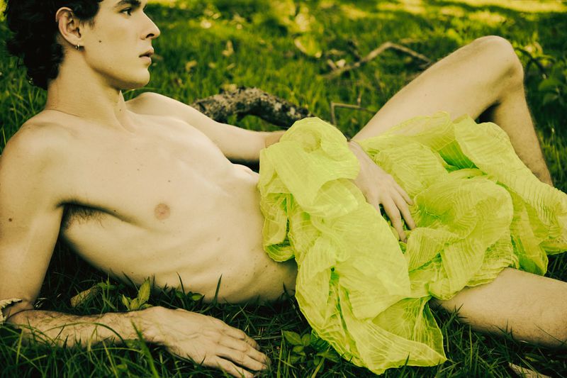 Nude adult male lies on grass with chartreuse textile. He is positioned outdoors in a natural environment with green foliage.