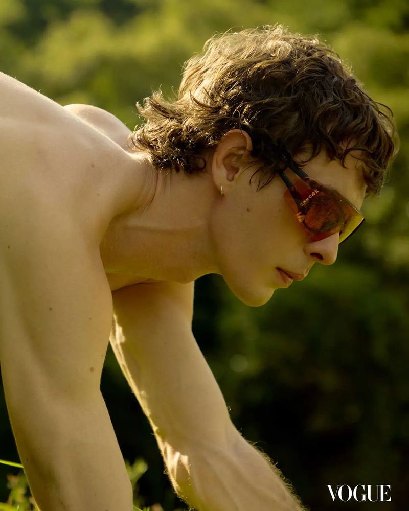 An adult male with curly brown hair and sunglasses is outdoors in a field of green. The man's torso is bare and in focus.