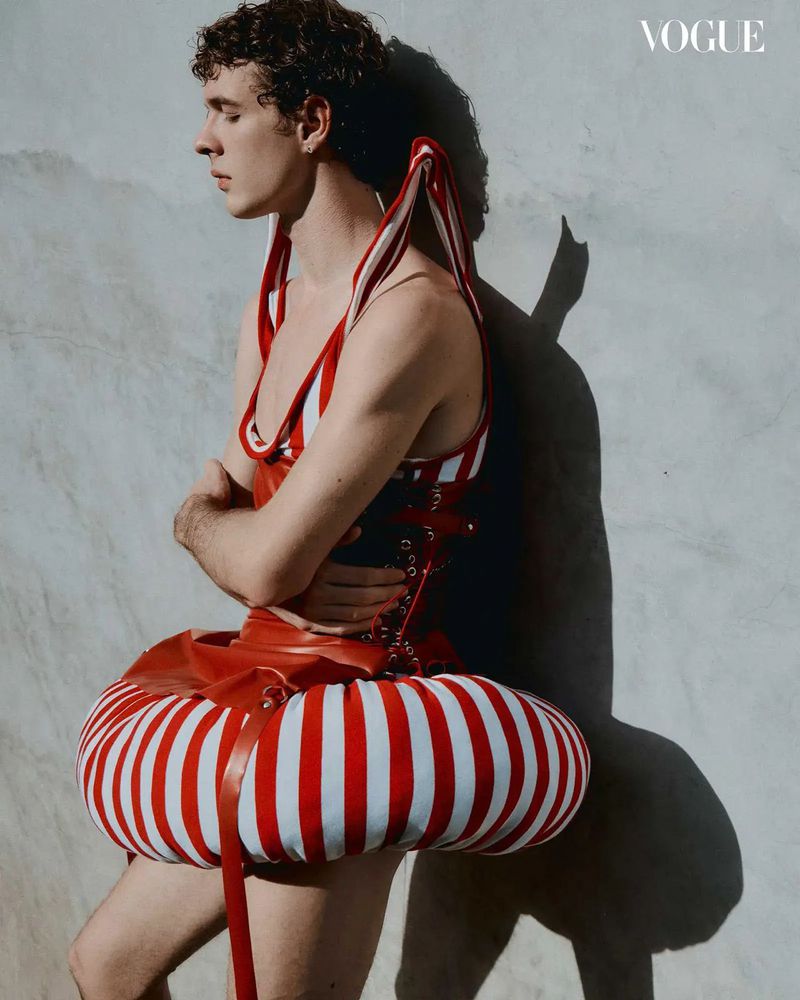 Fashion editorial shot of a male model in avant-garde striped red and white outfit, posing against a neutral background, with 'Vogue' logo visible.