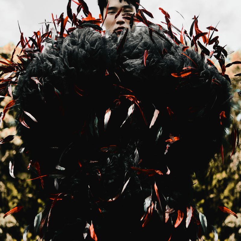 Fashion-forward portrait of an Asian male model wearing a black feathered couture creation, set against a soft-focus natural background.