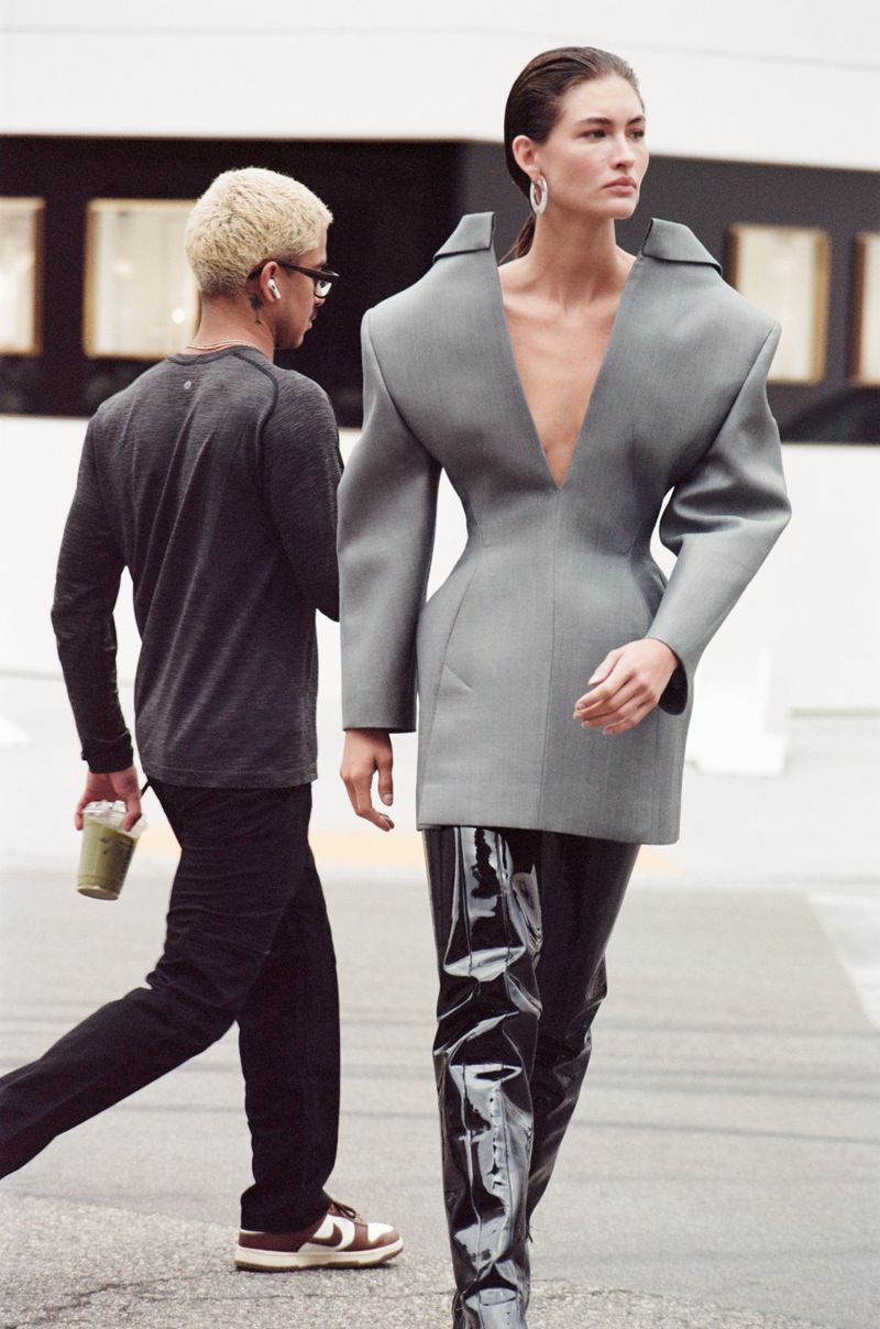 Fashionable woman in oversized grey blazer and thigh-high shiny boots walks on a city street with a man carrying a drink.