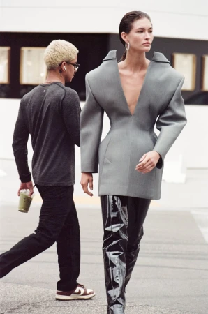 Fashionable woman in oversized grey blazer and thigh-high shiny boots walks on a city street with a man carrying a drink.
