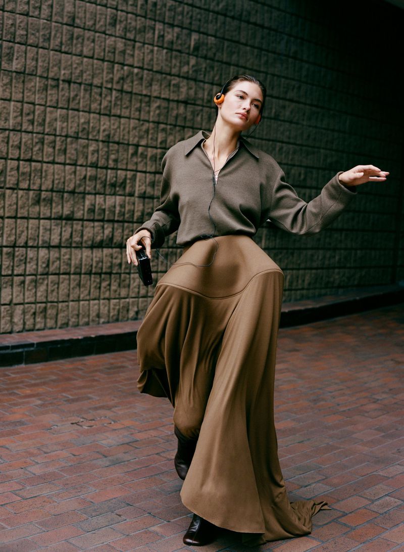 A fashionable young adult woman in a beige blouse and flowing skirt dances on a brick walkway with a brick wall in the background.