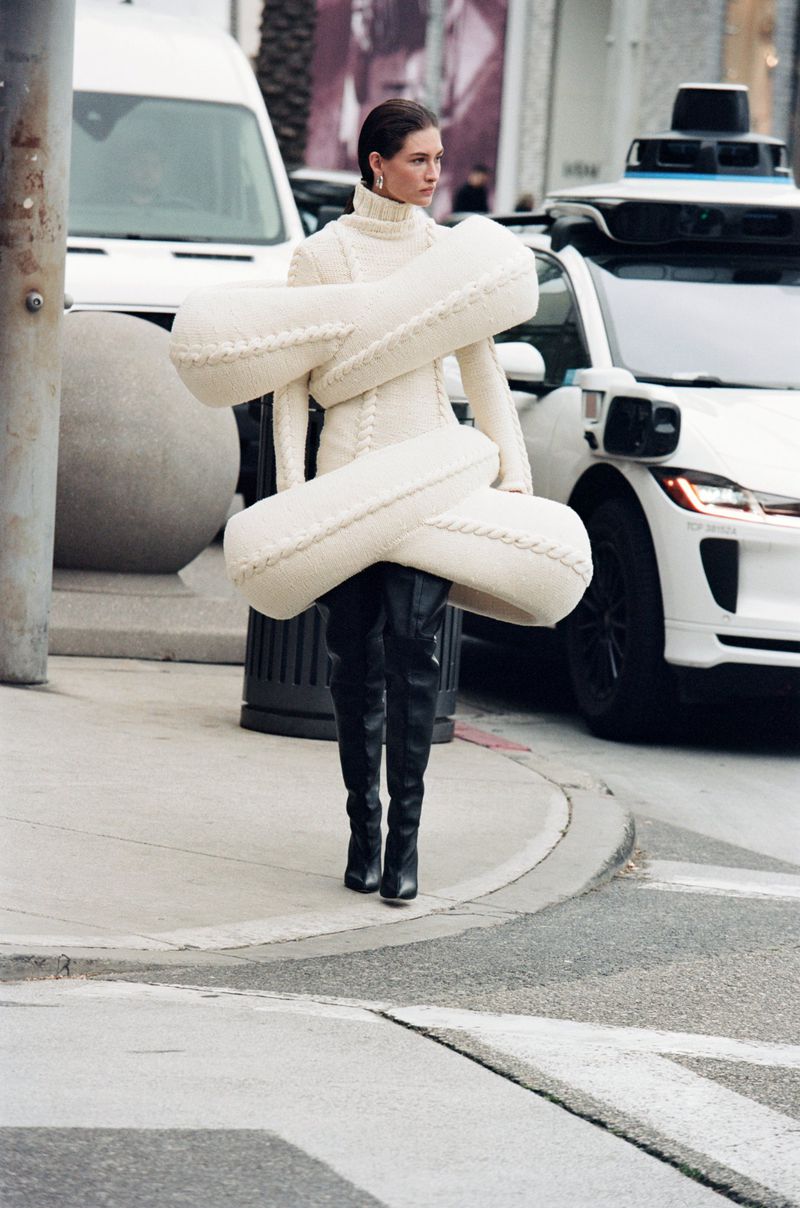 A fashion model poses on a city street wearing a cream-colored knit dress with oversized sculptural padding and black leather boots.
