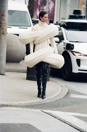 A fashion model poses on a city street wearing a cream-colored knit dress with oversized sculptural padding and black leather boots.