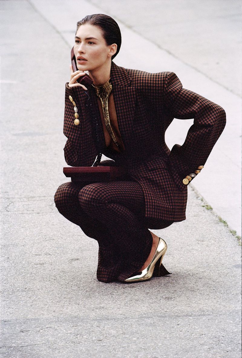 Stylish woman in houndstooth suit and gold heels crouches on asphalt using landline phone. Outdoor fashion editorial shot.