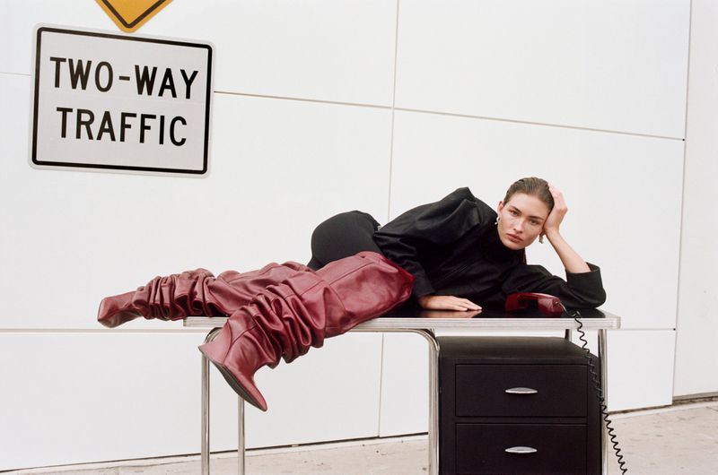 A woman in a black outfit is lying on a desk next to a telephone and wearing tall, slouchy red leather boots in a studio setting.