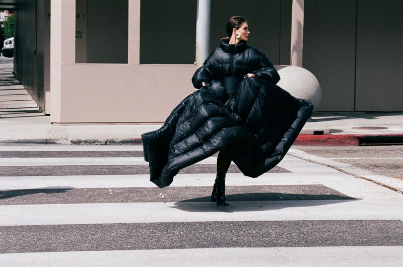 Adult woman in a large black puffer coat on a crosswalk in an urban setting. Neutral background, medium shot, looking off to the side.