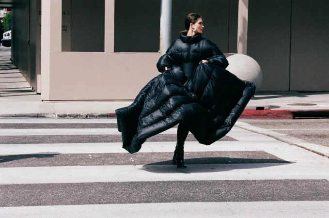 Adult woman in a large black puffer coat on a crosswalk in an urban setting. Neutral background, medium shot, looking off to the side.