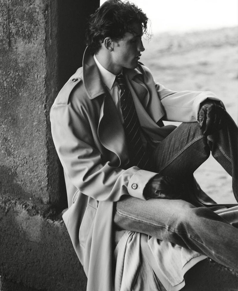 Model wearing a trench coat, striped tie, and denim jeans seated near a concrete wall opening in black and white.