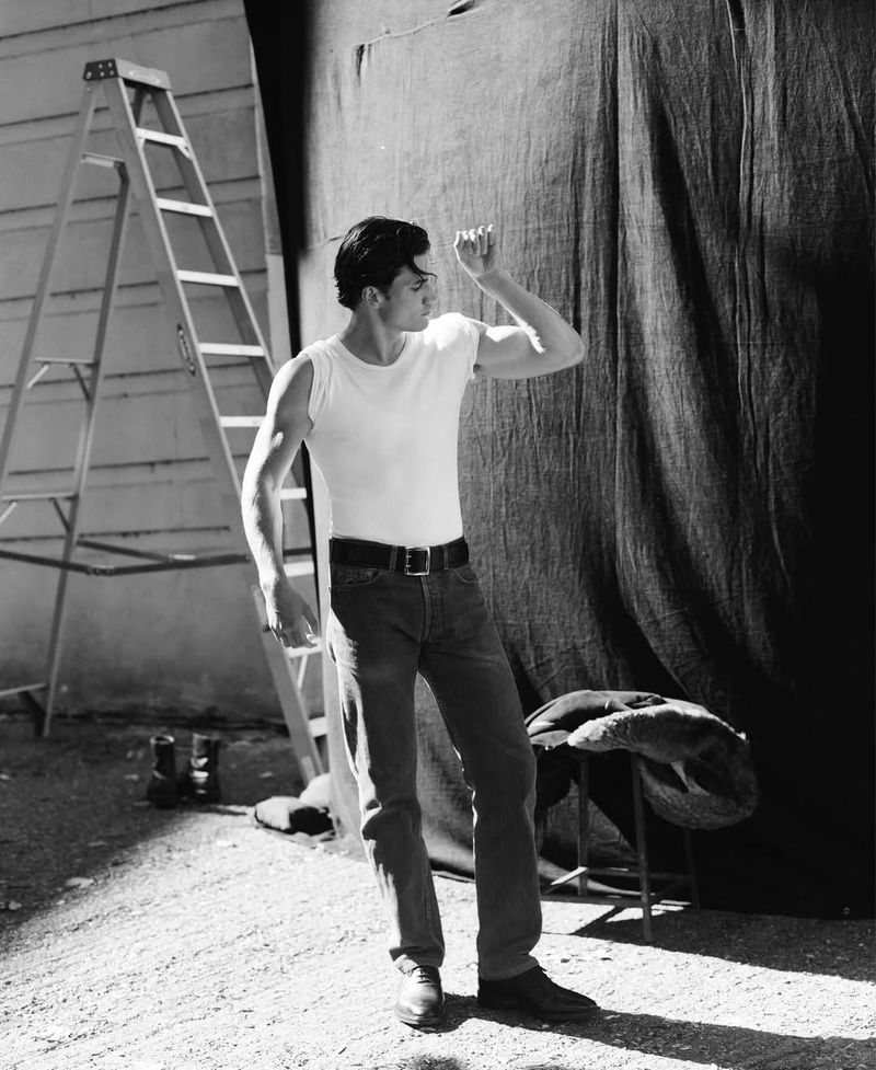 Monochromatic shot of a man in a white tank top and jeans, standing in an outdoor setting with a ladder and draped fabric backdrop.