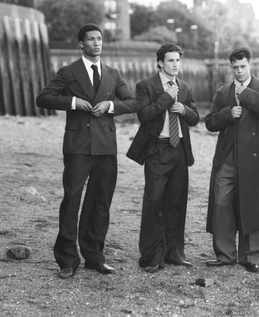 Three male fashion models pose outdoors, wearing stylish suits and ties in a black-and-white fashion editorial shot near a harbor.