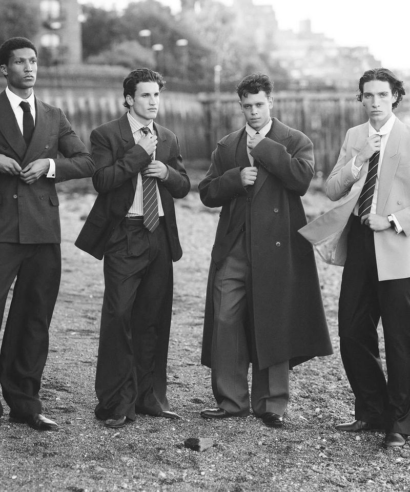 Four men in suits stand outdoors against a blurred background. The men are adjusting ties or jackets. A monochrome image, full shot.