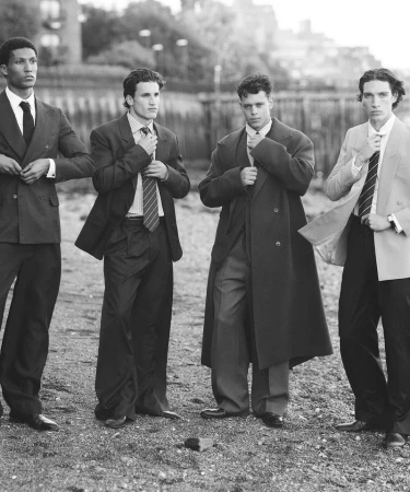 Four men in suits stand outdoors against a blurred background. The men are adjusting ties or jackets. A monochrome image, full shot.