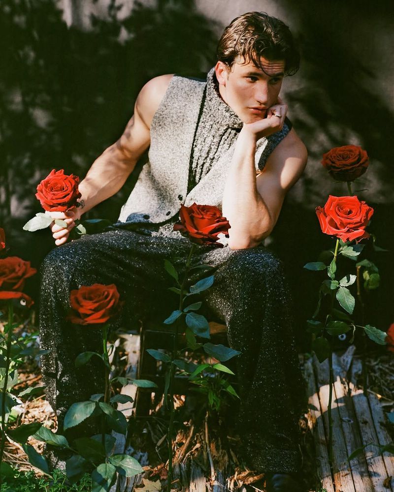 Handsome man in a grey vest seated among red roses in a garden setting. He rests his chin in his hand, creating a stylish pose.