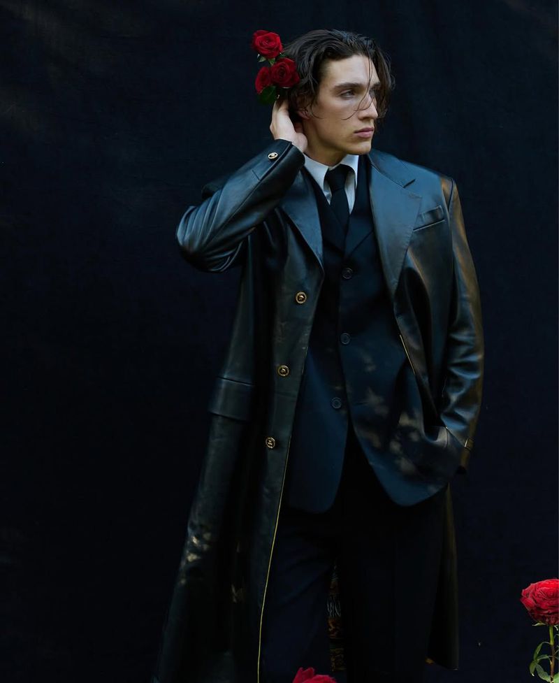 A stylish, young adult male model wearing a black leather coat, vest, and trousers, complemented by red roses against a dark backdrop.