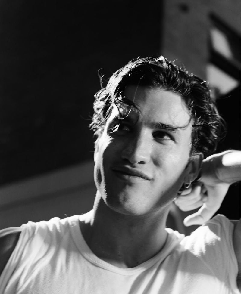 Monochrome portrait of a young adult man with wet curly hair, wearing a white tank top, looking up in a low-key studio setting.