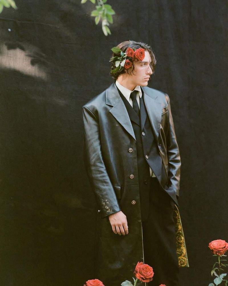 Adult male in a black coat and floral headpiece posing in front of a dark backdrop, with red roses scattered near the bottom of the frame.