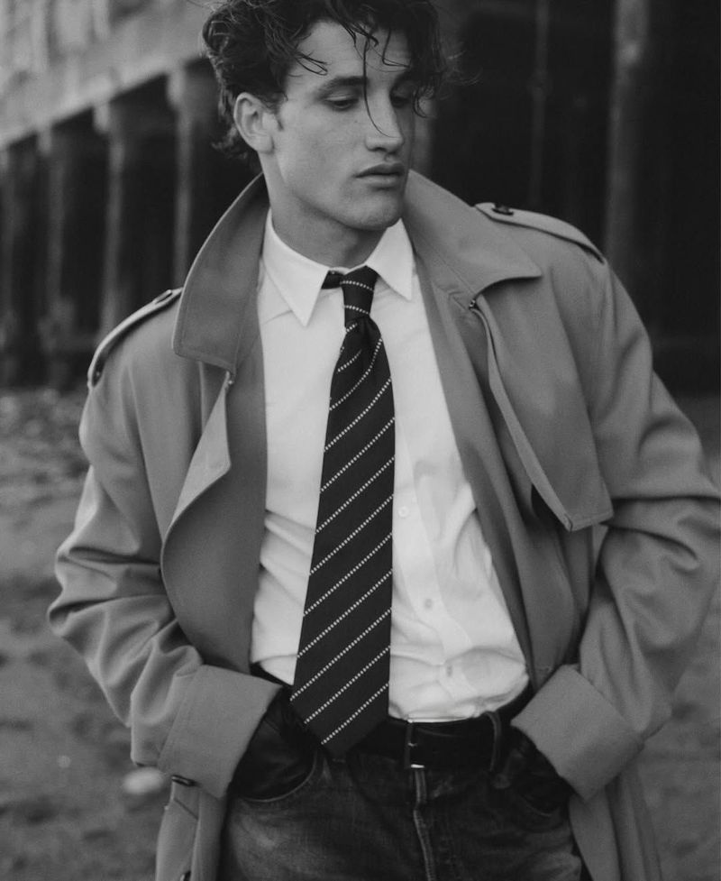 Black and white portrait of a young adult man in a trench coat, white shirt, and tie, standing outdoors with a serious expression.
