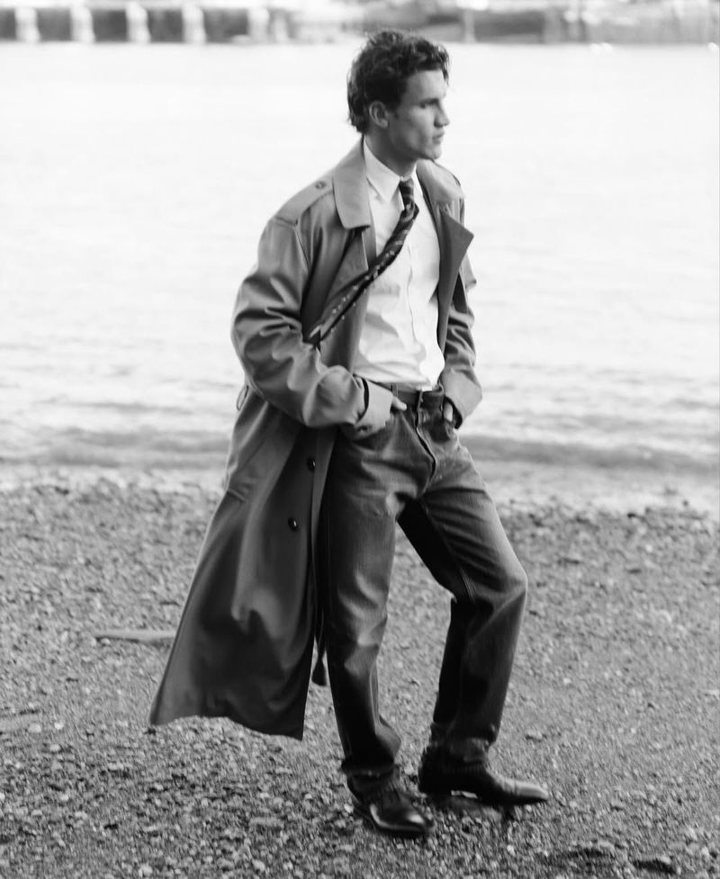Monochrome image of an adult male in a trench coat standing on a rocky beach near calm water, looking away from the camera.