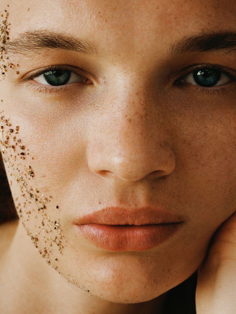Close-up portrait of an adult woman with light skin and freckles, and light-brown decorative beads applied to her cheek and forehead.