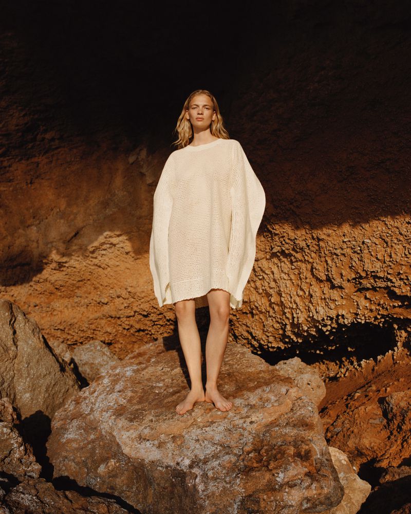 Adult woman standing barefoot on a rock wearing an oversized cream-colored knit dress in a rocky outdoor environment.