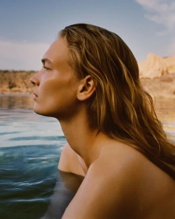 Close-up side profile of a woman with wet hair wading in calm coastal water under warm natural sunlight.