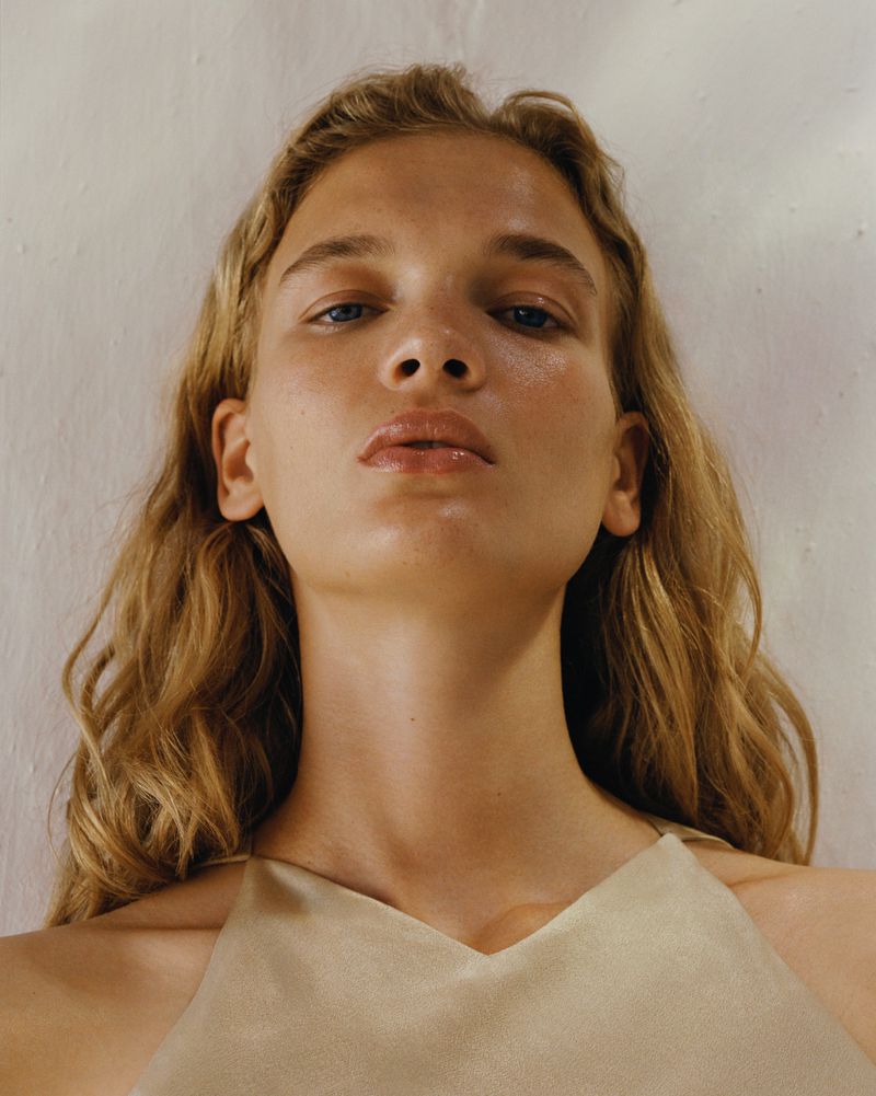 Close-up portrait of a young blonde woman with a neutral expression. She wears a simple cream-colored top against a textured white wall.