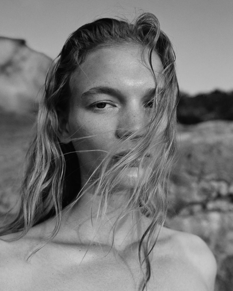 Black and white close-up portrait of model Caitlin Soetendal with windblown hair over her face against a rocky background.