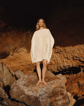 Model Caitlin Soetendal standing barefoot on a rock wearing an oversized cream knit poncho dress in direct sunlight.