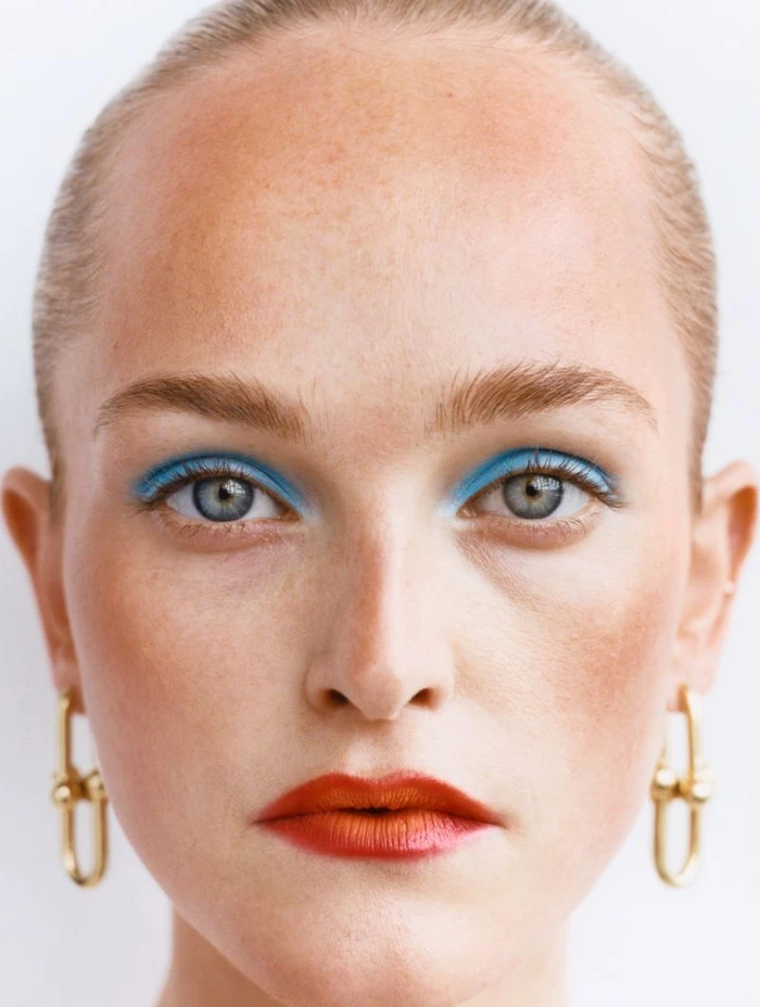 Close-up portrait of a woman with vibrant blue eyeshadow, red lipstick, slicked hair, and gold link earrings against white seamless