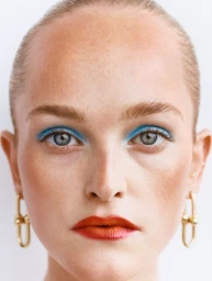 Close-up portrait of a woman with vibrant blue eyeshadow, red lipstick, slicked hair, and gold link earrings against white seamless