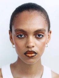 Close-up beauty portrait of a woman with slicked hair, dewy skin, gold hoop earrings, and glossy metallic ombré lipstick.